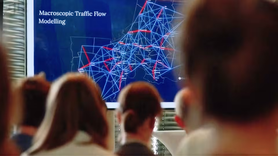 Audience watching presentation on macroscopic traffic flow modelling with map of Europe overlaid by blue and red routes