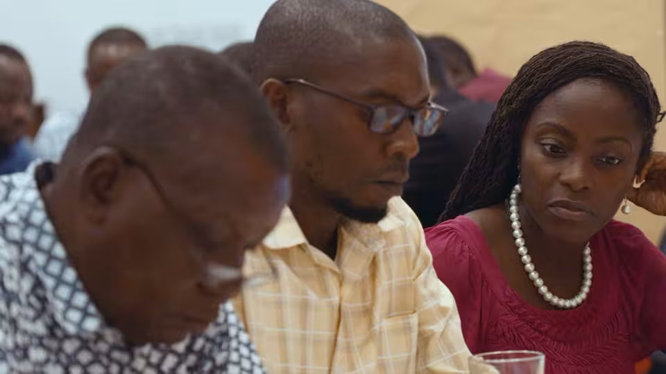 Three adults sit close together in workshop setting, two men focused on notes, woman with pearl necklace listening