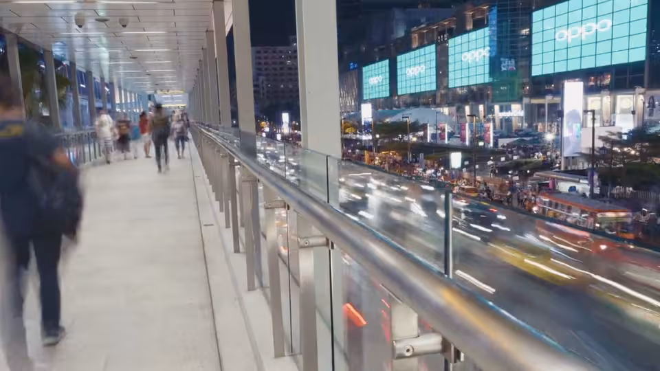 Pedestrian skywalk with evening commuters, below a busy city road with blurred car lights and bright digital billboards