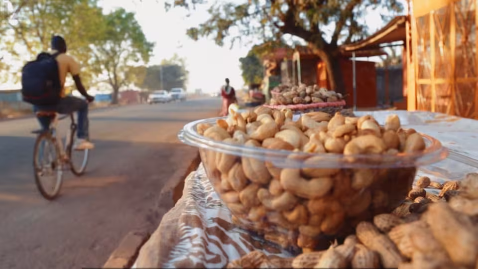 Plastic bowl of cashew nuts on roadside stall in focus, with cyclist and pedestrians moving along sunlit rural street