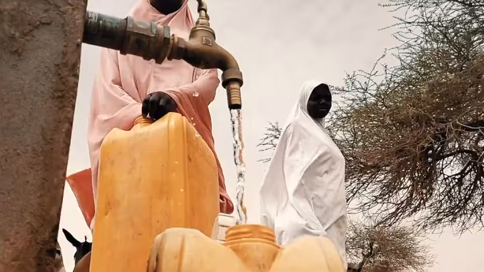 People filling large yellow water canisters at an outdoor tap, with a woman in a white headscarf standing nearby.
