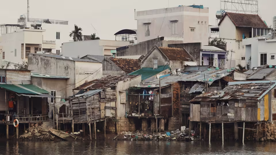 Densely packed wooden stilt houses and small buildings along a riverbank with modern structures in the background.