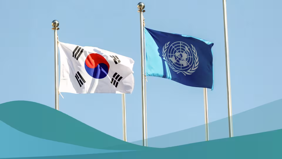 The national flag of South Korea and the flag of the United Nations fly side-by-side on metal flagpoles against a clear blue sky, with a teal wave graphic across the bottom.