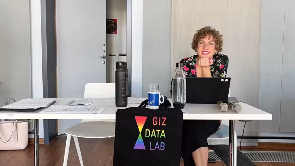 A woman with curly brown hair sits behind a desk, smiling at the camera, with a black laptop in front of her. In the foreground, a black bag hangs off the table, featuring a rainbow prism logo and the text "GIZ DATA LAB" in multicolored letters. Multiple papers, two water bottles, two microphones, and a mug are arranged on the desk.